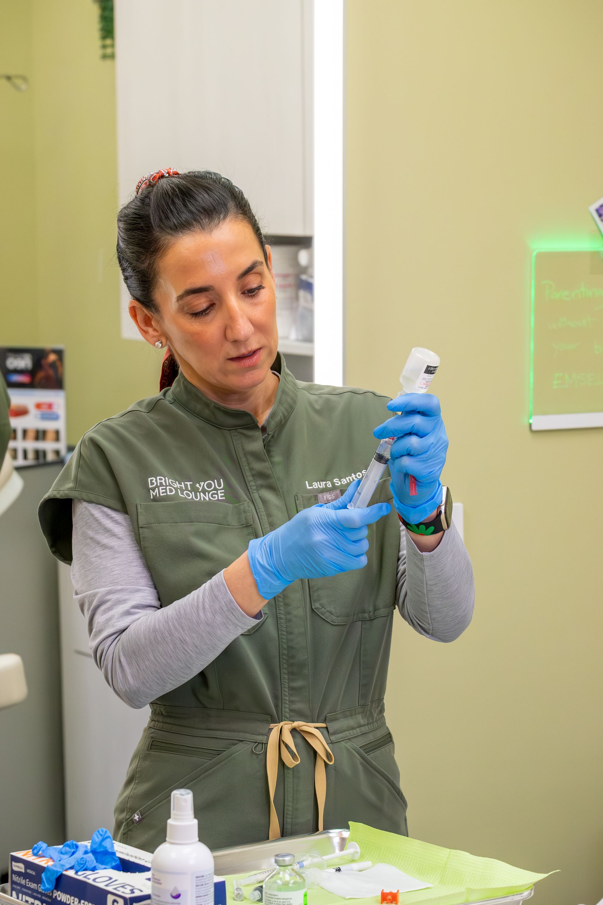 Healthcare worker preparing a syringe in a clinical room, wearing gloves and scrubs.