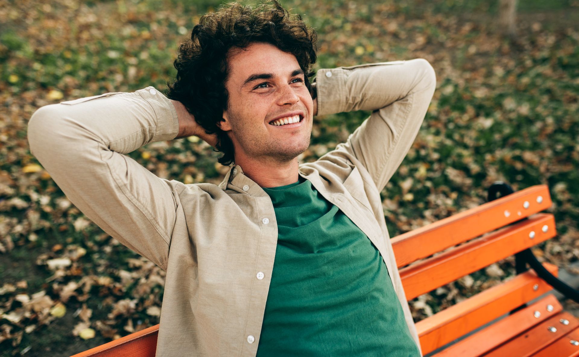 Smiling person reclining on an orange bench in a park, hands behind head, wearing a beige shirt and green T-shirt