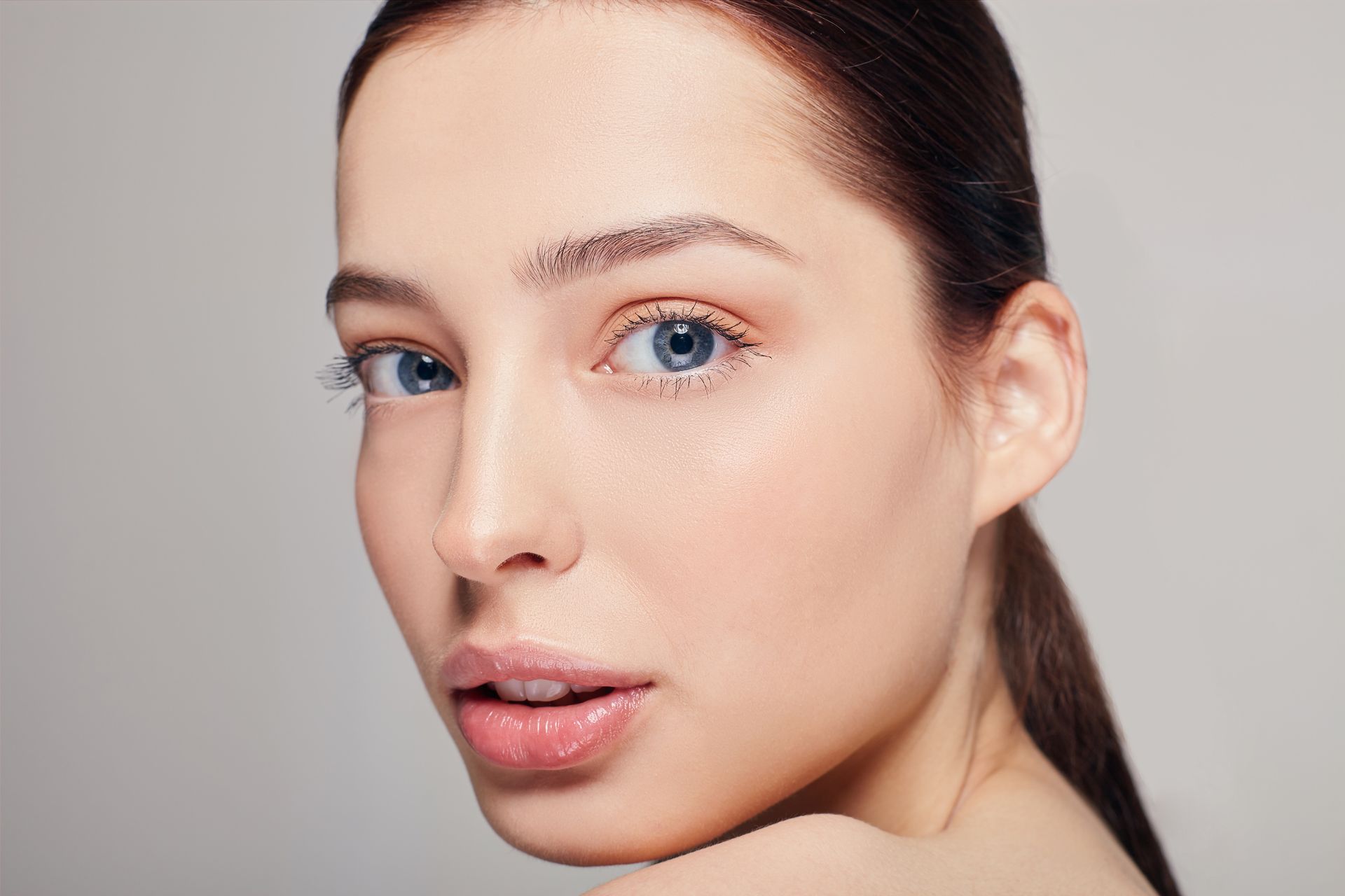Close-up of a woman with blue eyes and natural makeup, looking over her shoulder against a light background