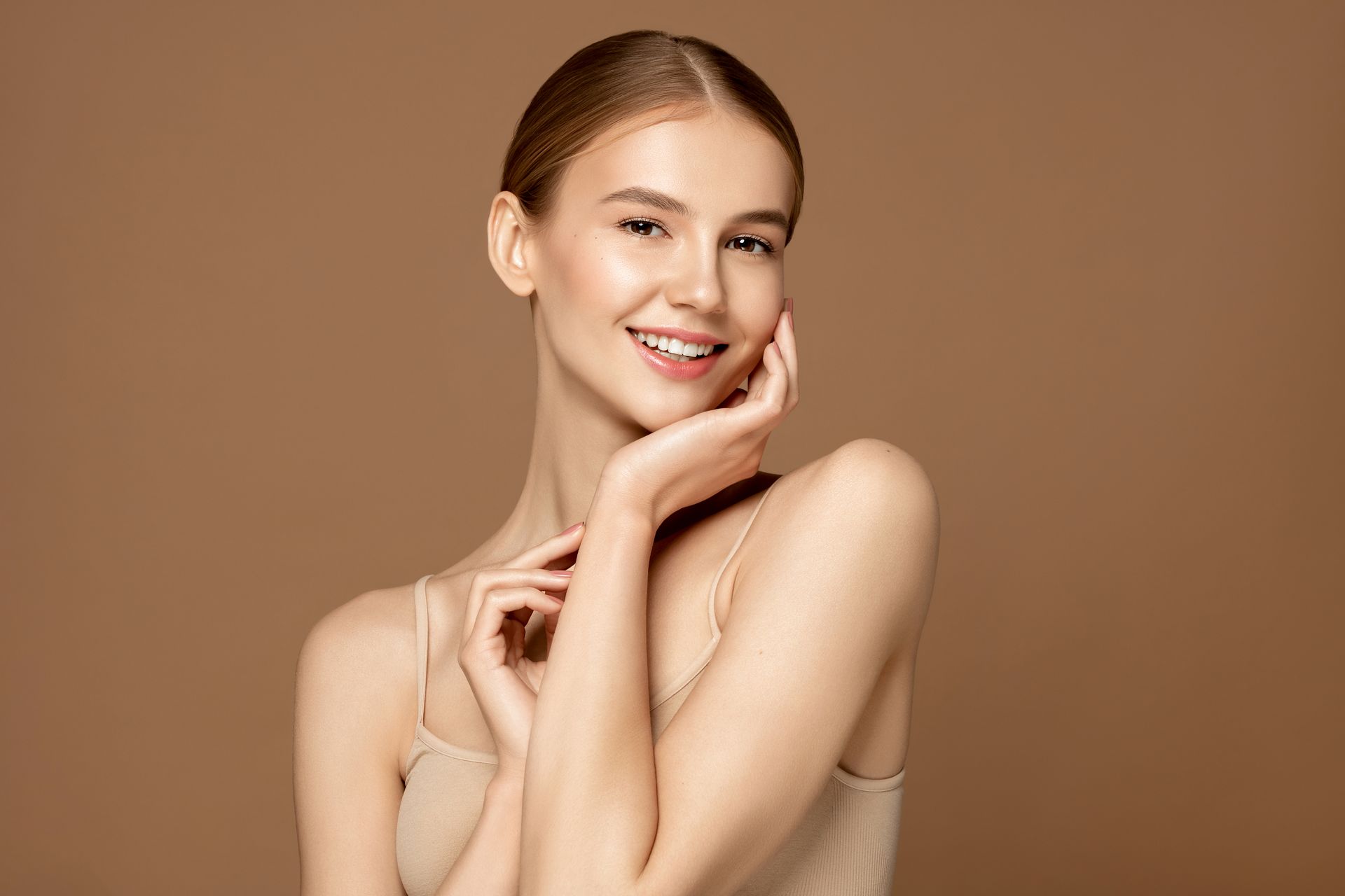 Woman smiling, hands near face, wearing a neutral-colored tank top, against a brown background.