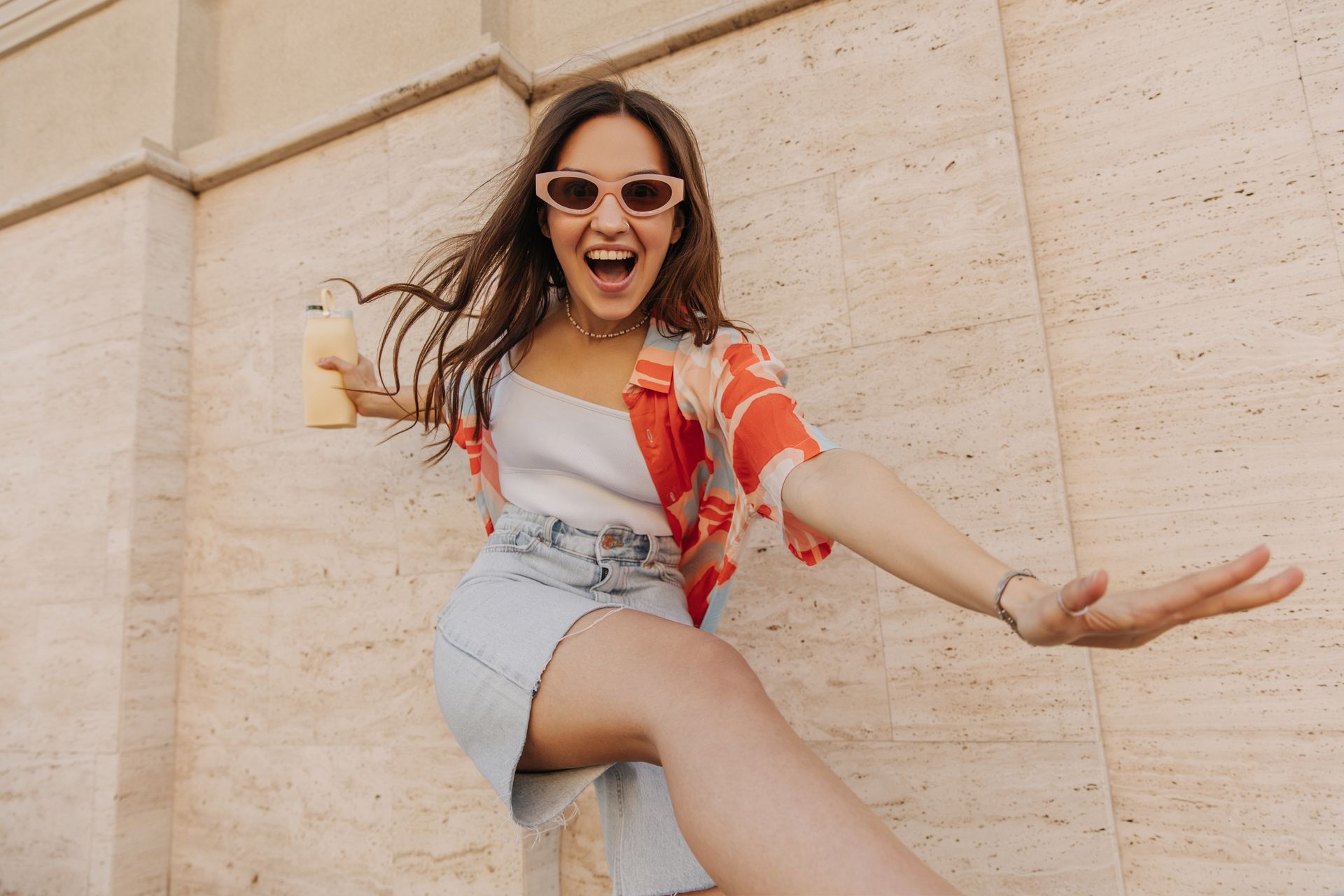 Woman in sunglasses, open-mouthed, holds drink, extends arm.  She wears a skirt and patterned top against a beige wall.