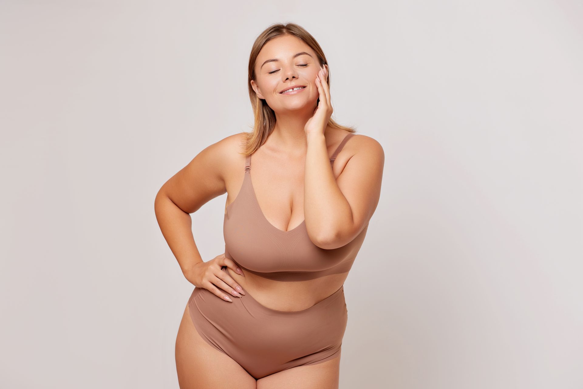 Woman posing in beige lingerie against a light background, hand on hip and smiling gently
