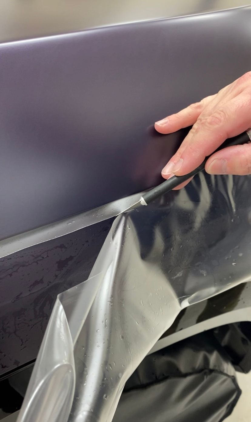 Person peeling protective film from a dark car panel.
