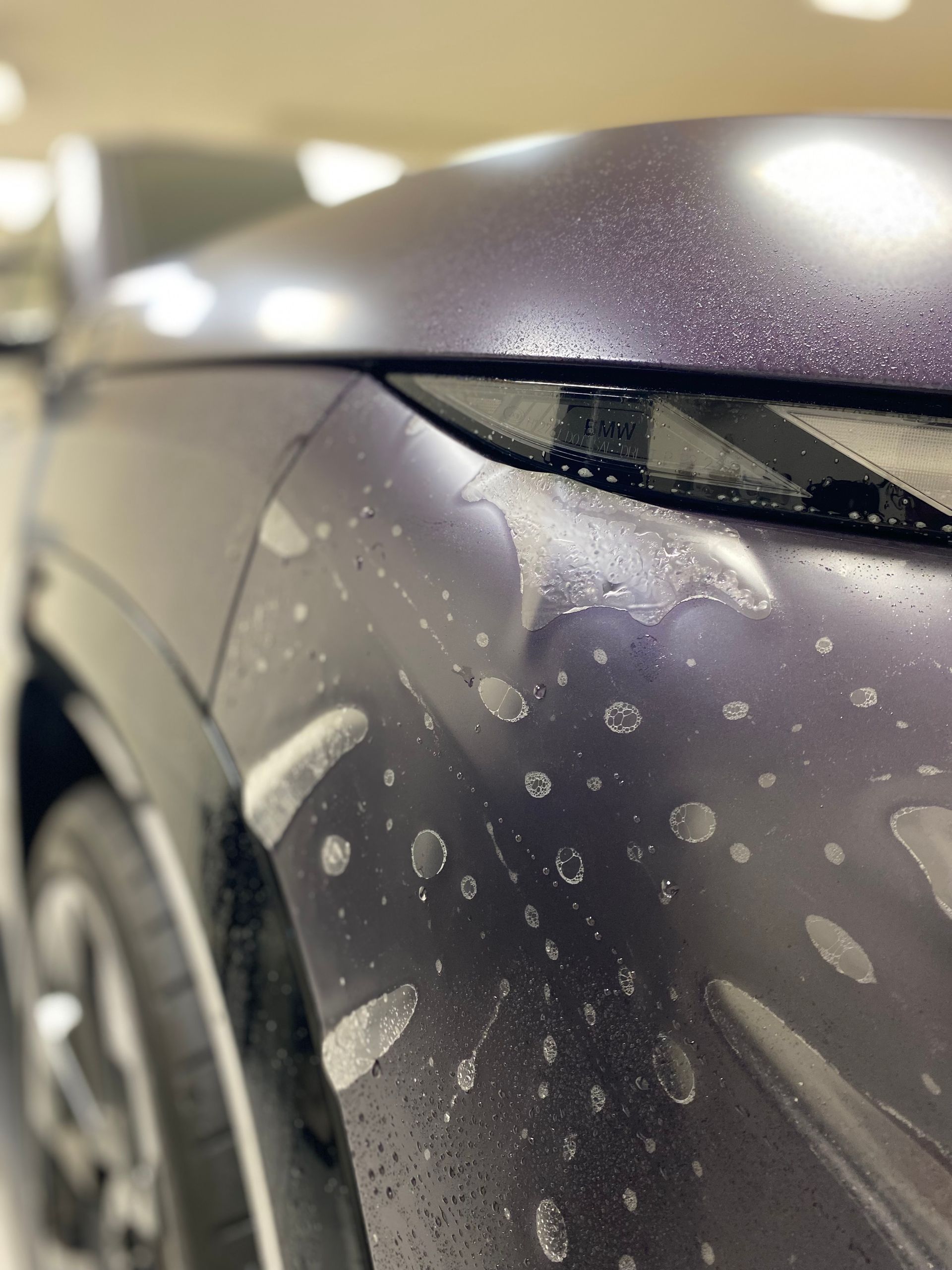 Dark blue car fender with water spots. Close-up, the car's headlight and wheel visible.