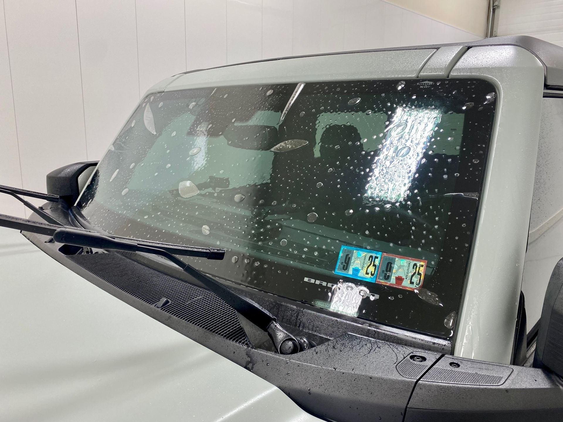 A car's windshield with water droplets; wiper blades are visible. Inside the car, a license plate is seen.