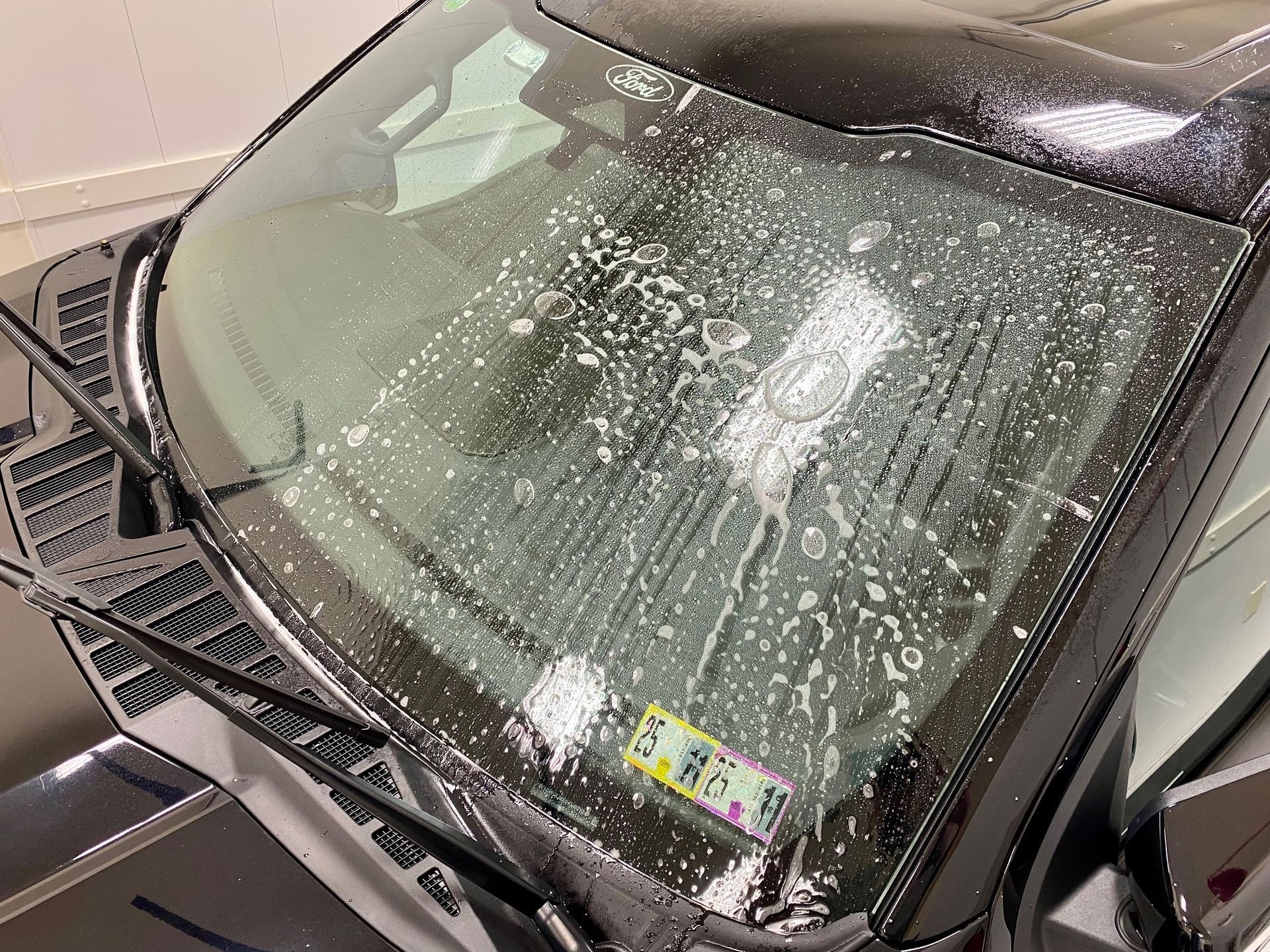 Black car windshield covered in soapy water. Yellow and green sticker visible.