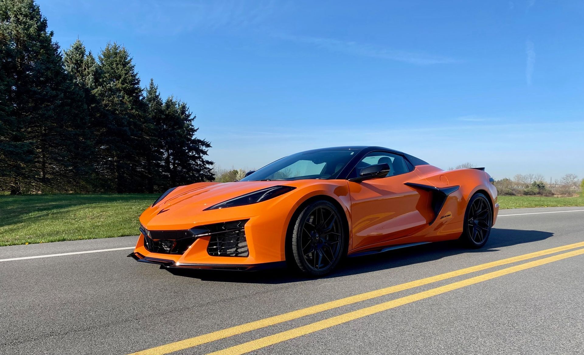 Orange sports car on a road, black roof, parked in front of trees under blue sky.