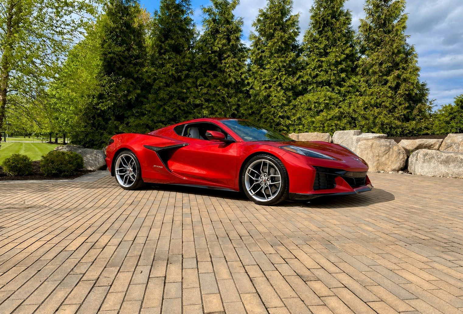Red sports car parked on a brick driveway beside trees and stone walls under a blue sky