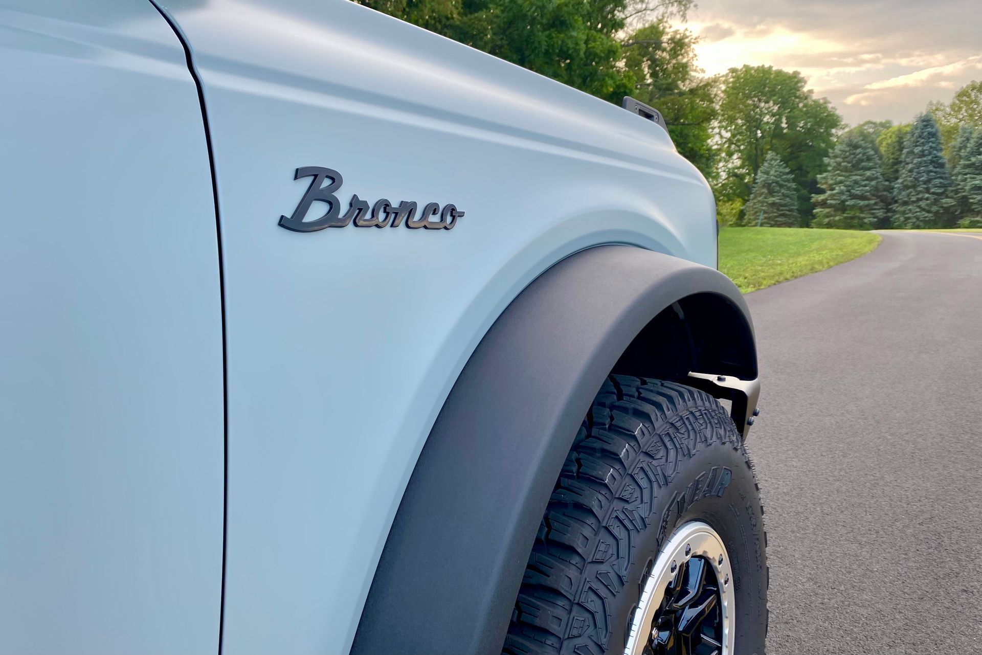 Light blue Ford Bronco, black fender, and tire on a road with trees.