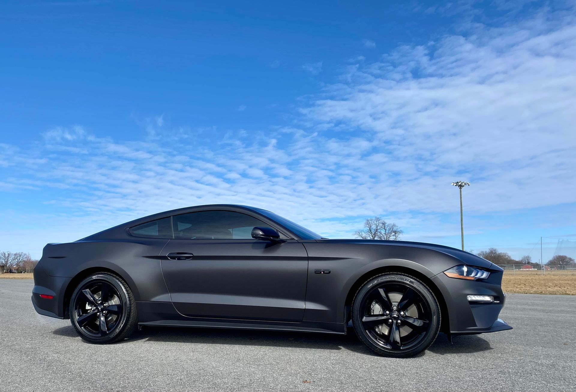 Dark gray Ford Mustang coupe with black wheels, parked on asphalt, bright blue sky.