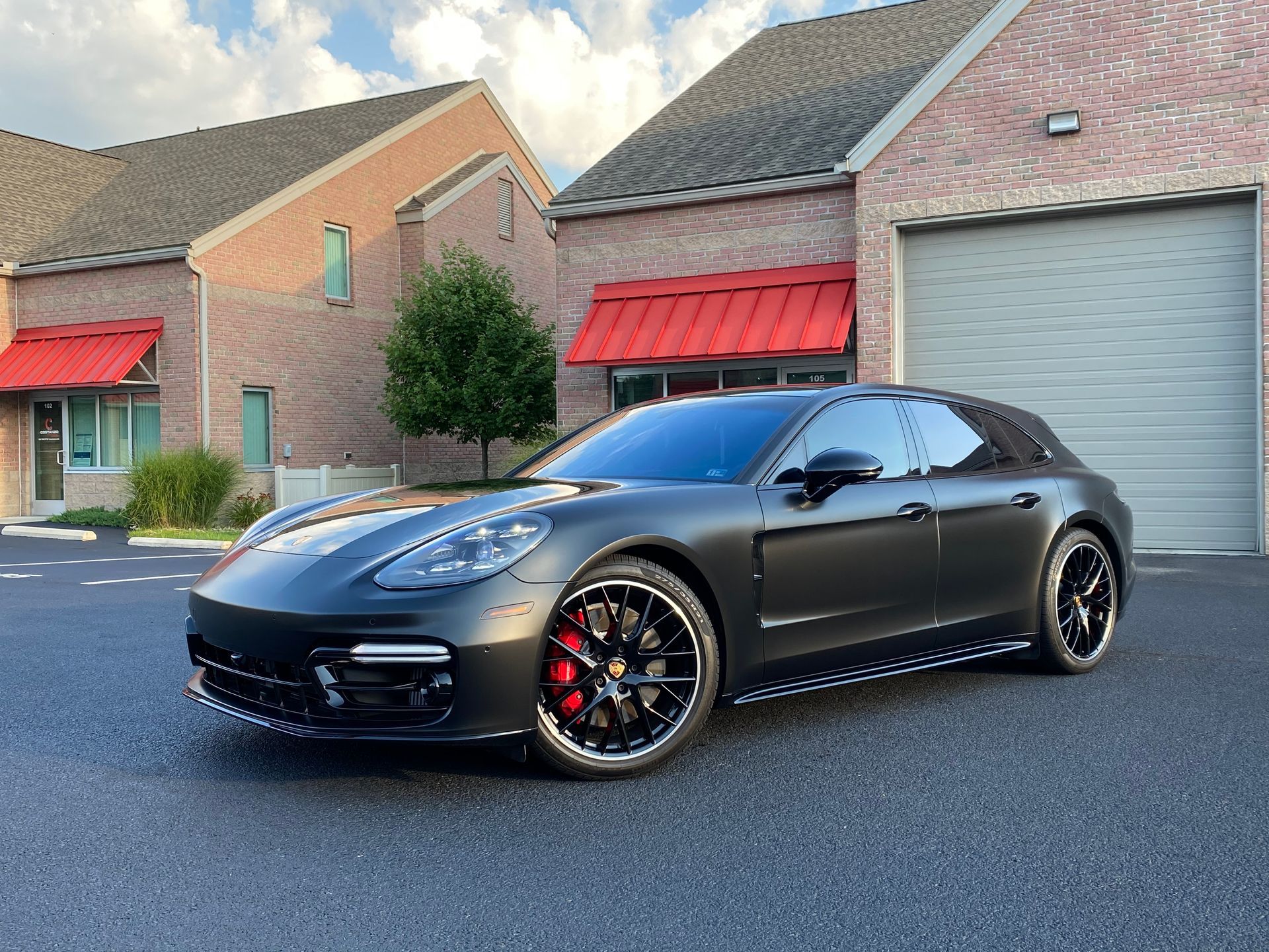 Dark gray Porsche Panamera parked in front of a brick building with red awnings and a garage door.