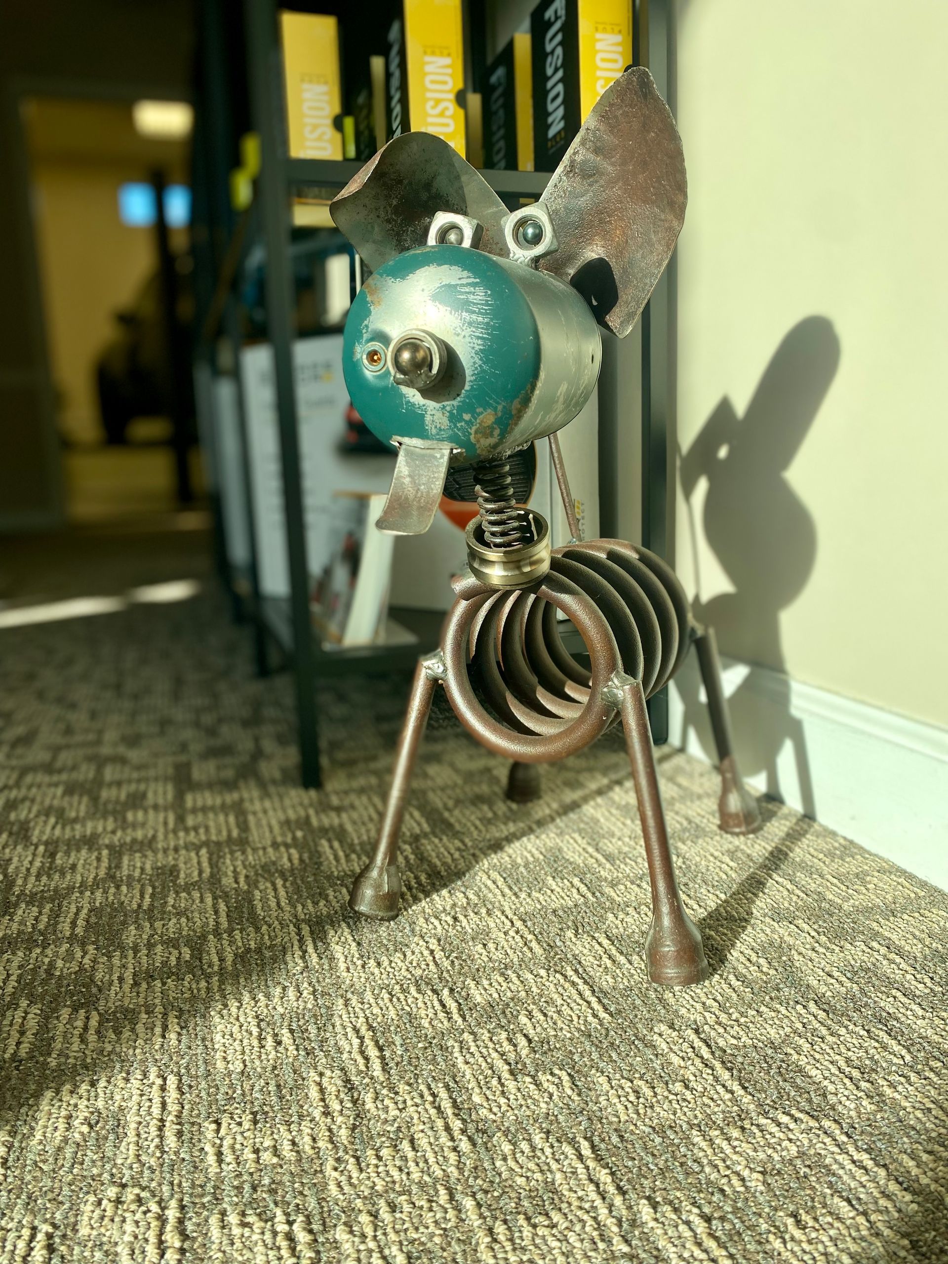 Metal dog sculpture with a teal head stands on a carpeted floor in a hallway.