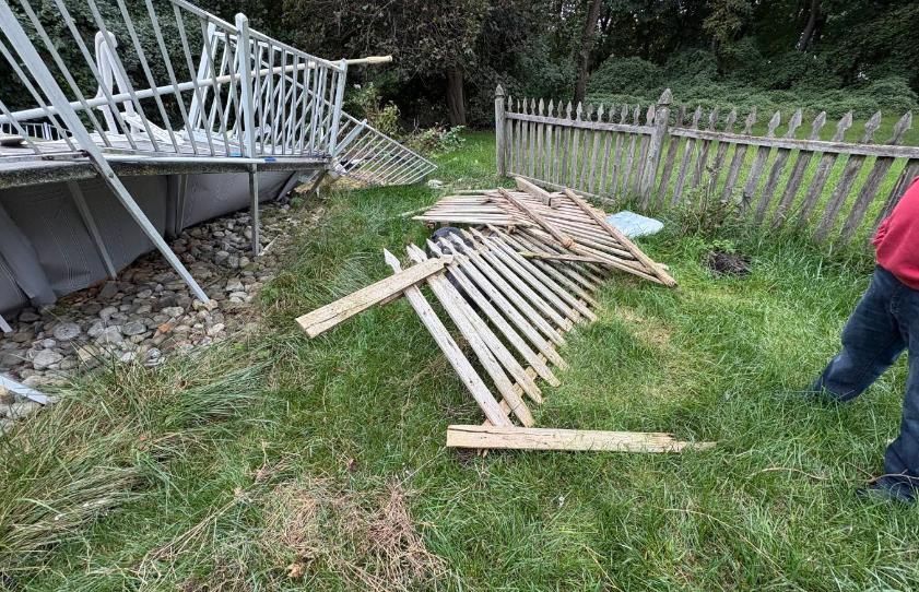 Broken wooden fence panels on grass beside a raised deck, with a person standing at the right edge