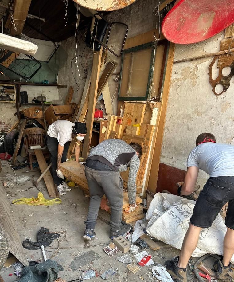 People cleaning debris in a damaged, cluttered room with broken walls and scattered rubble.