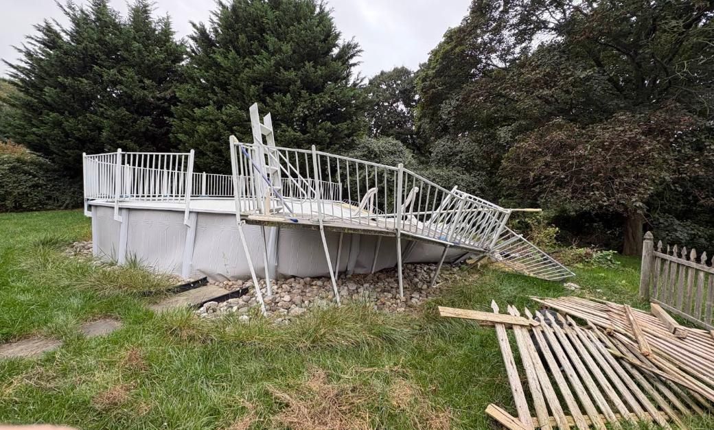 Broken white deck railing collapsed in a grassy yard beside trees and a fence.