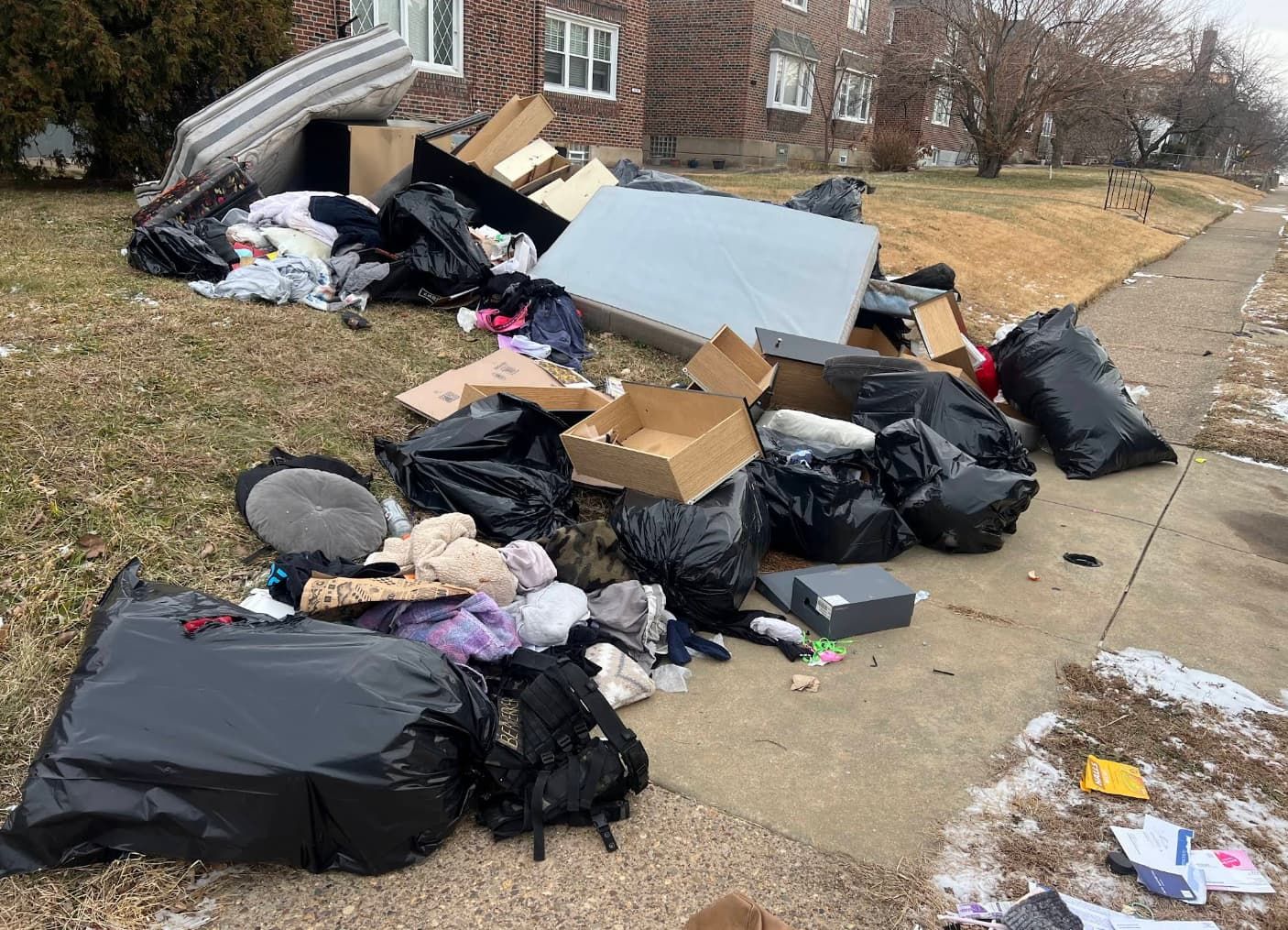 Overflowing trash bags and scattered debris piled along a sidewalk beside apartment buildings.