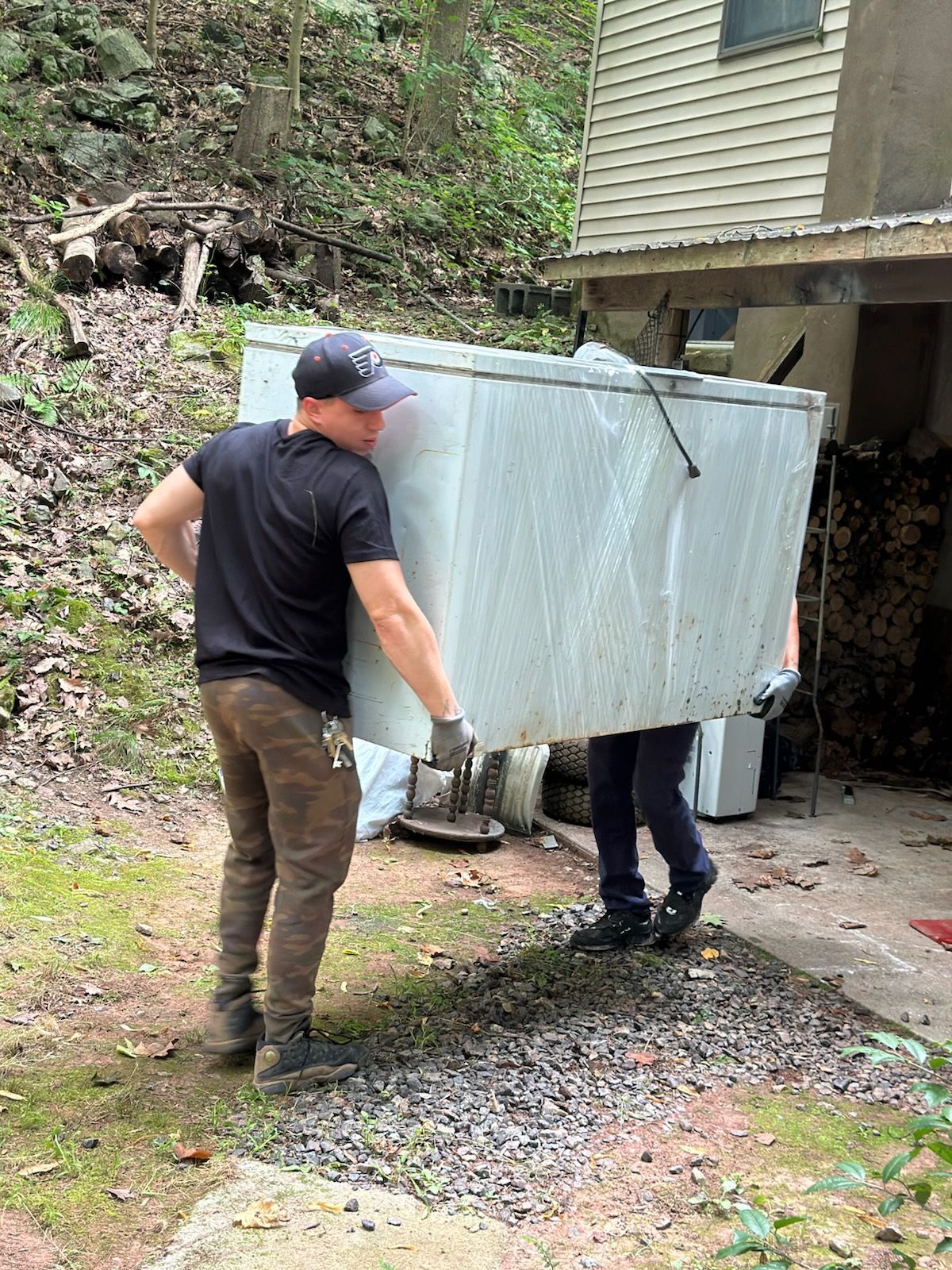 Two people carrying a large wrapped appliance up a gravel driveway beside a house.
