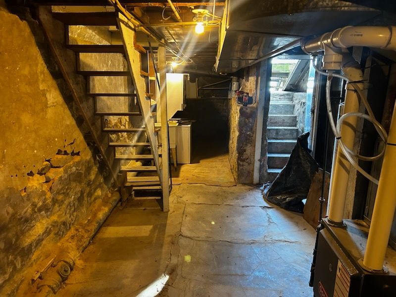 Dim basement stairway with metal steps, concrete floor, yellow walls, and exposed pipes and wiring