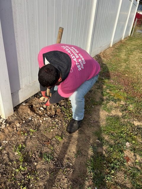 Person in pink shirt crouches by a fence, digging in soil near a building.