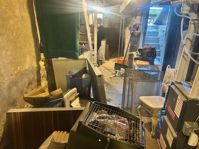 Narrow basement utility room with tools, shelves, bins, and a grated machine under bright overhead lights