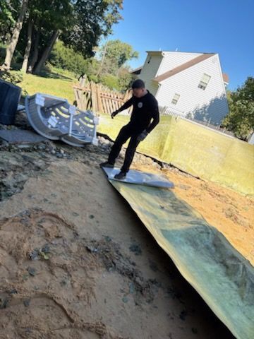 Person standing on a tarp-covered dirt yard beside construction materials and a wheelbarrow, near a white house.