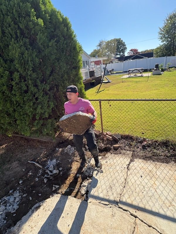 Person carrying a large dirt clump beside a chain-link fence in a sunny yard