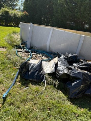 Black yard bags and a blue hose beside a white above-ground pool in a grassy yard