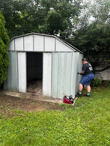 Person painting a small metal shed beside trees and grass.
