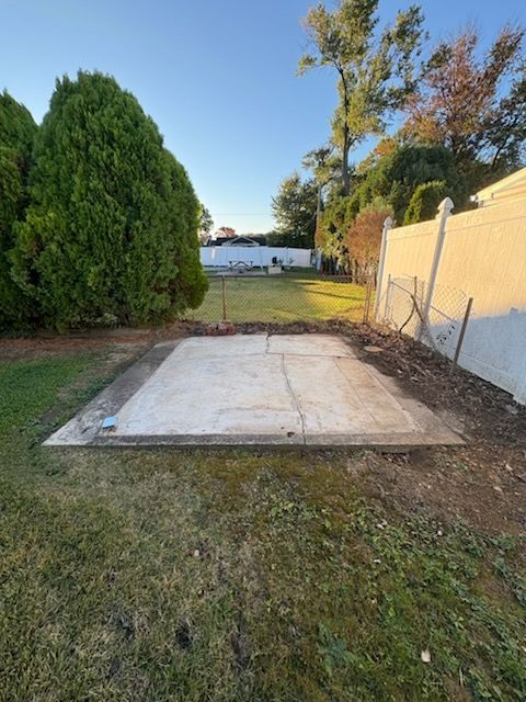Concrete slab in a backyard beside a fence, with grass, trees, and a house in the distance.