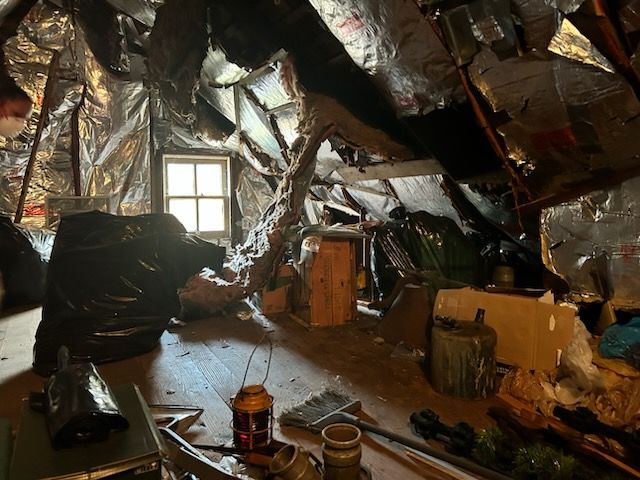 Dim attic with torn insulation, small window, and scattered boxes and tools on the floor.
