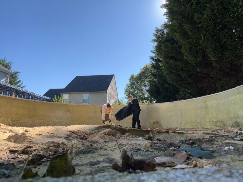 Two children play in a rocky, sand-filled backyard beside a tall fence under a blue sky.