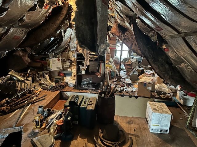 Cluttered attic with exposed beams, boxes, tools, and scattered debris under a dark roof