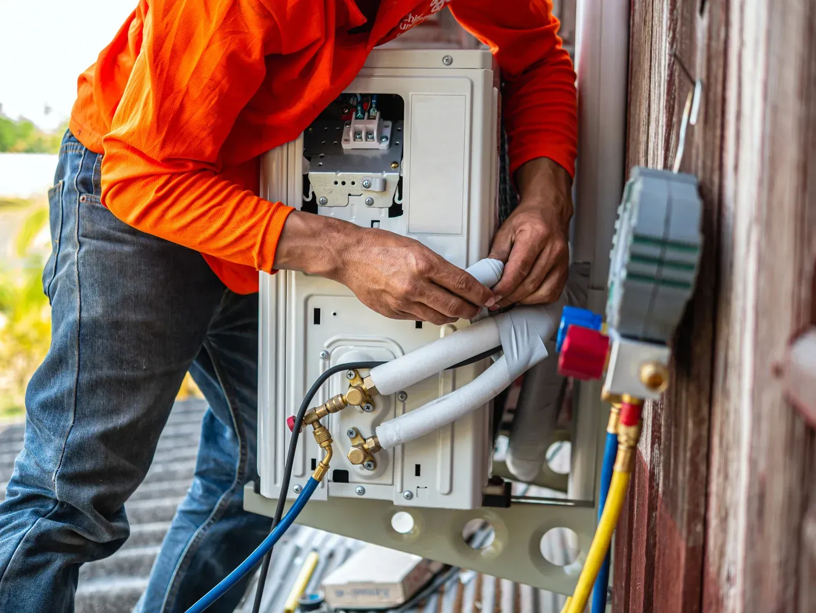 Person in orange shirt installing an air conditioning unit outside, connecting tubes.