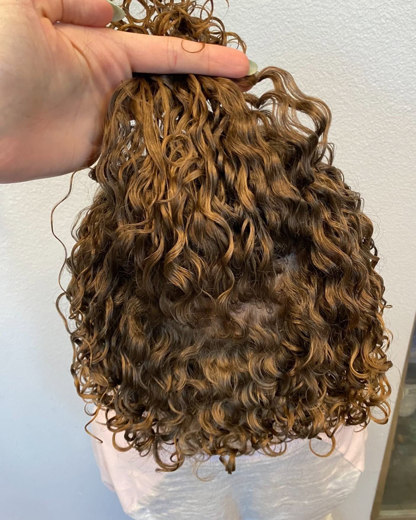 Hand holding a bundle of curly light brown hair extensions against a white background