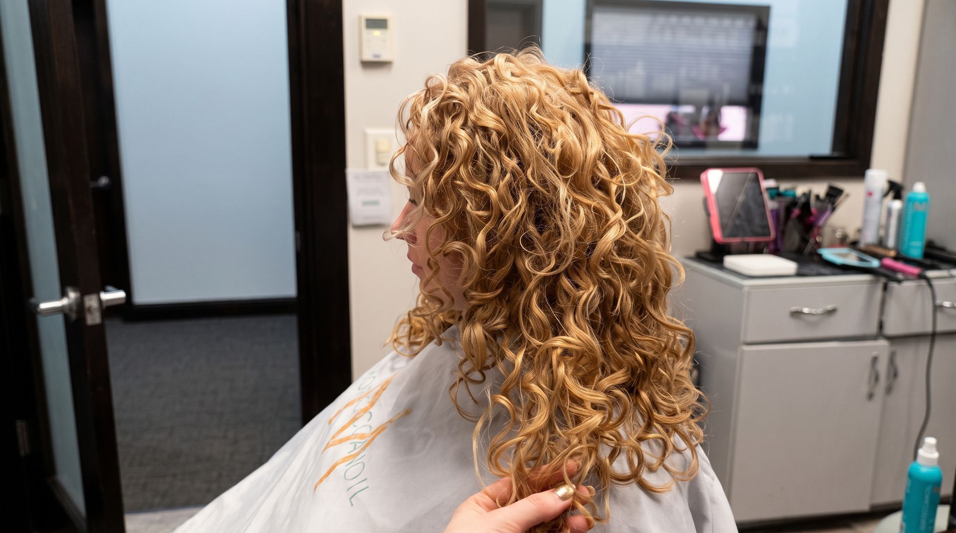 Person with long curly dark hair in a salon chair, shown in profile wearing a light cape.