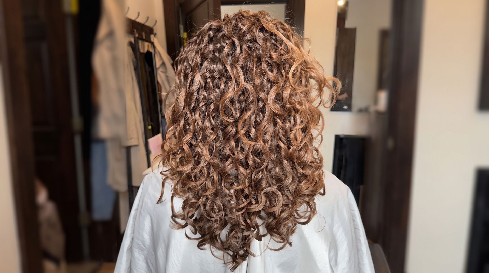 Long, curly dark hair falling down the back, with arms raised against a light wall.
