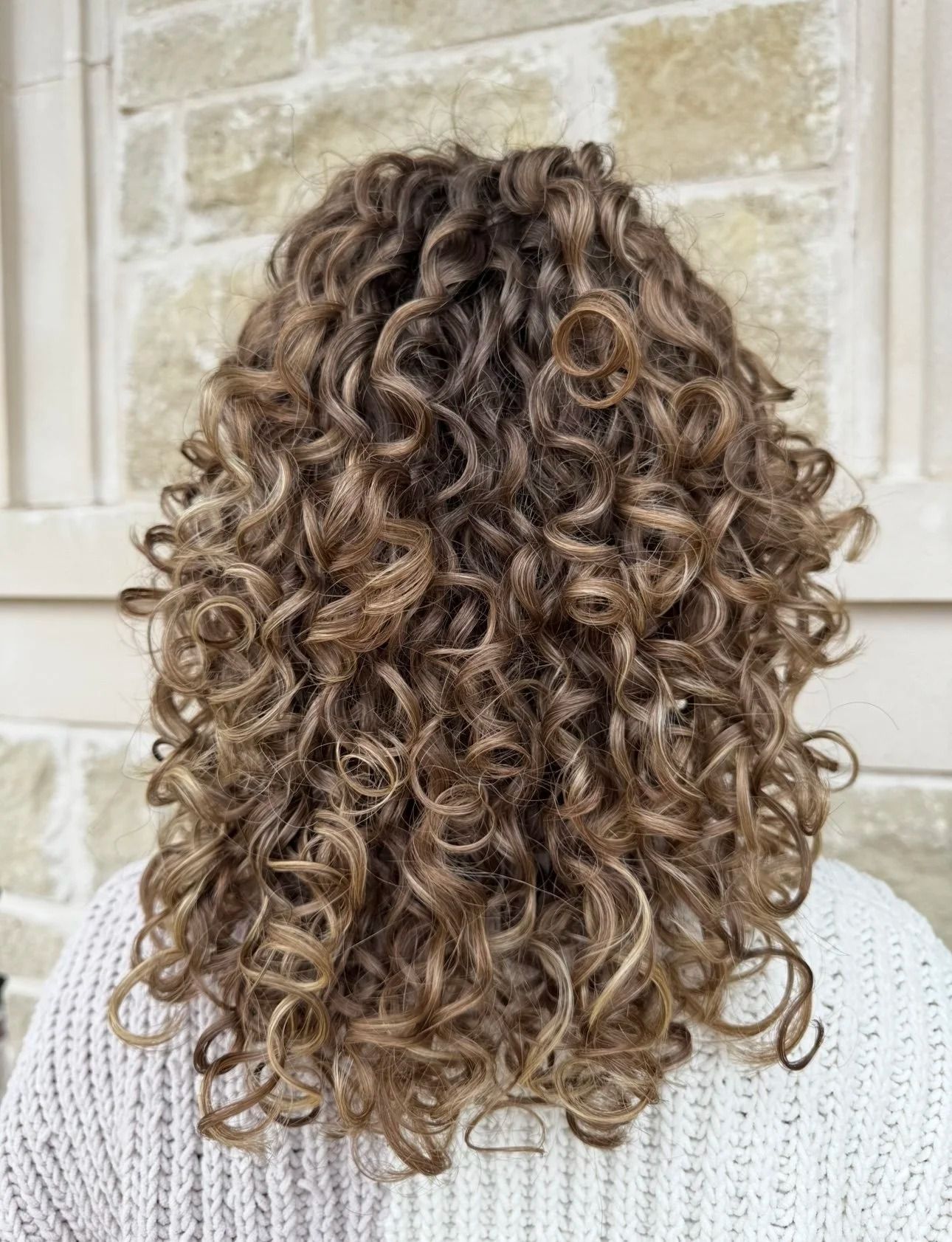 Back view of shoulder-length curly brown hair against a stone wall background