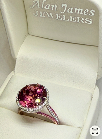 A large, oval pink gemstone ring with a diamond halo, displayed in an open Alan James Jewelers white ring box.