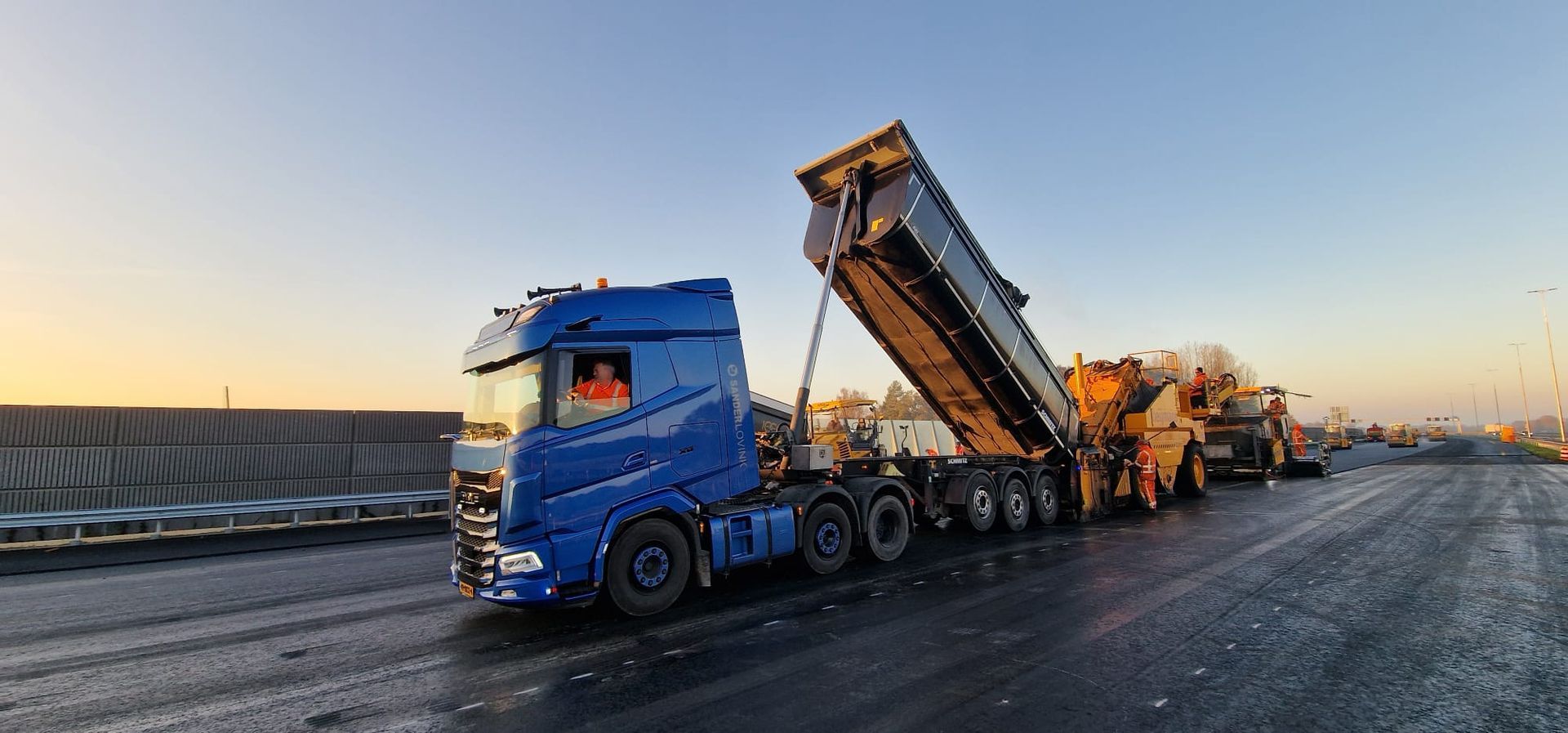 Een blauwe kiepwagen lost asfalt op een wegwerkterrein bij zonsondergang.
