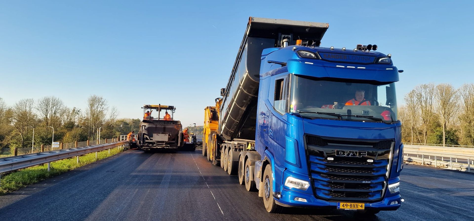 Blauwe kiepwagen en wegwerkvoertuigen op een geasfalteerde snelweg onder een heldere hemel.