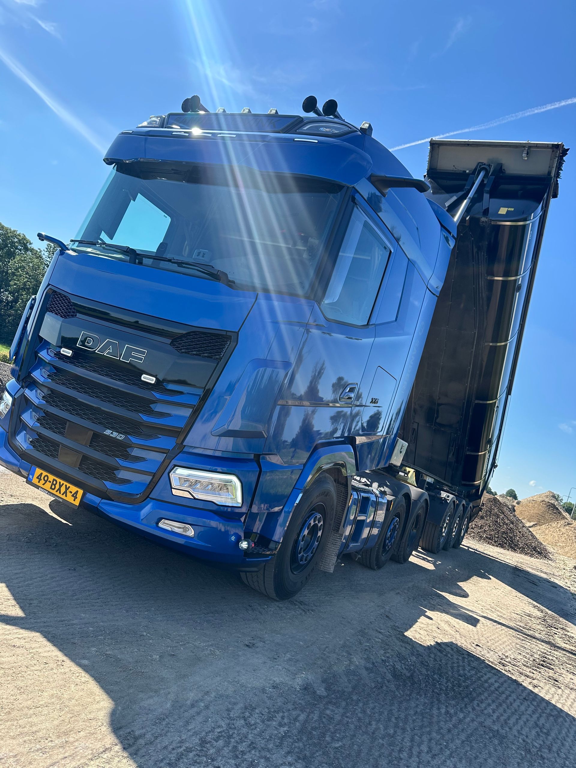 Blauwe kiepwagen met verhoogde laadbak geparkeerd op een grindterrein onder een heldere hemel.