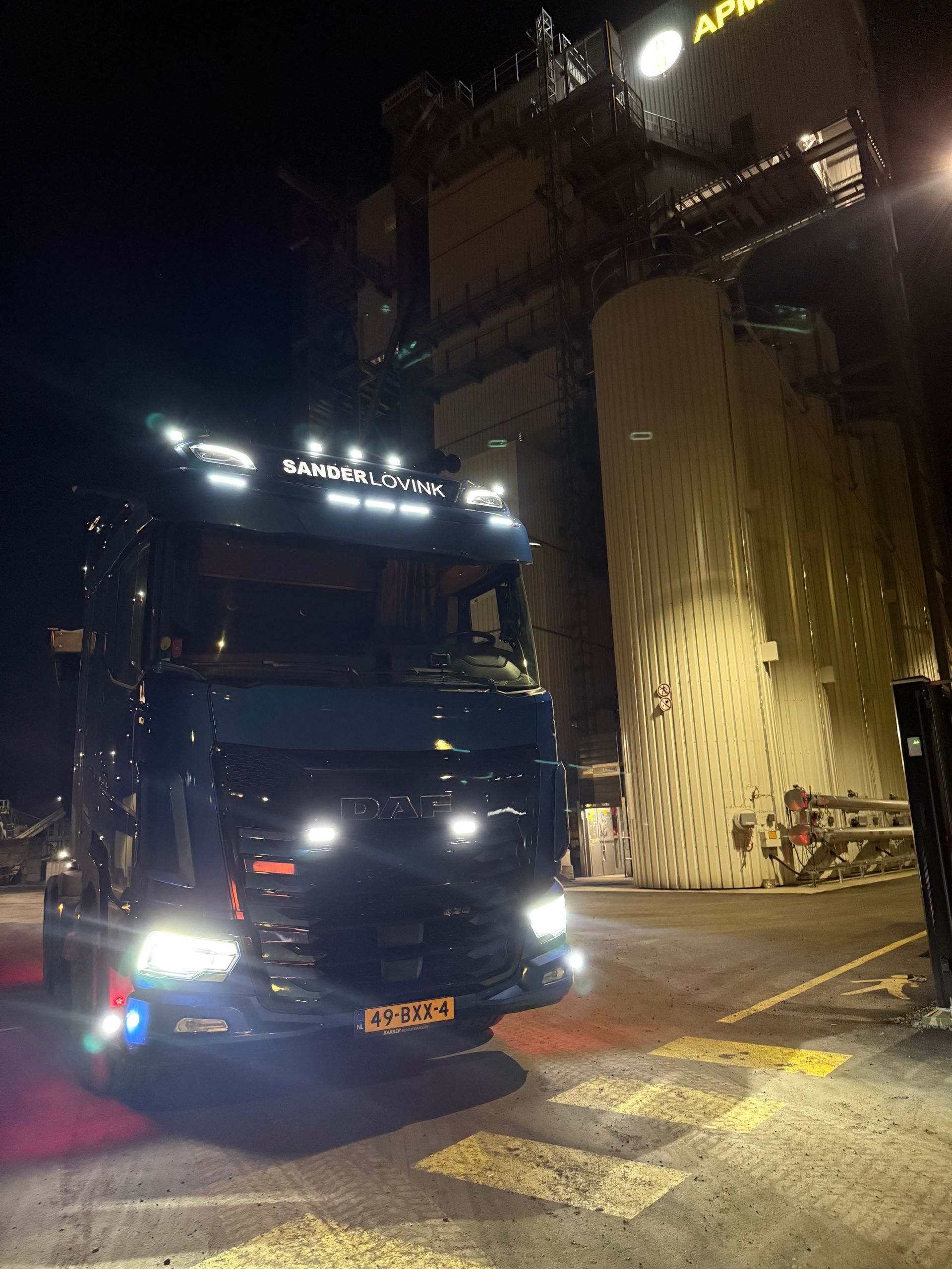 Zwarte vrachtwagen met felle koplampen 's nachts naast een hoog industriegebouw en een zebrapad.