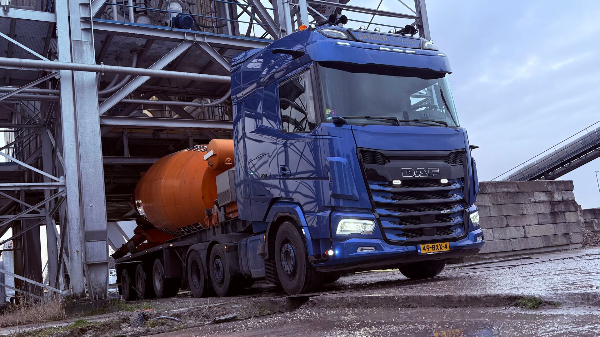 Een blauwe vrachtwagen met een oranje cementmixer rijdt door een industrieel bouwterrein.