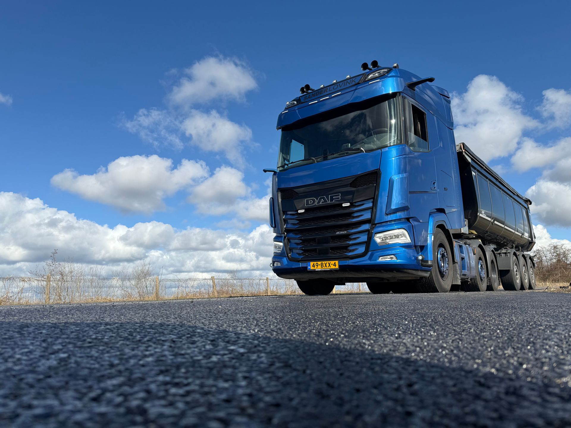 Een blauwe kiepwagen rijdt over een weg onder een bewolkte hemel, gezien vanuit een laag perspectief.