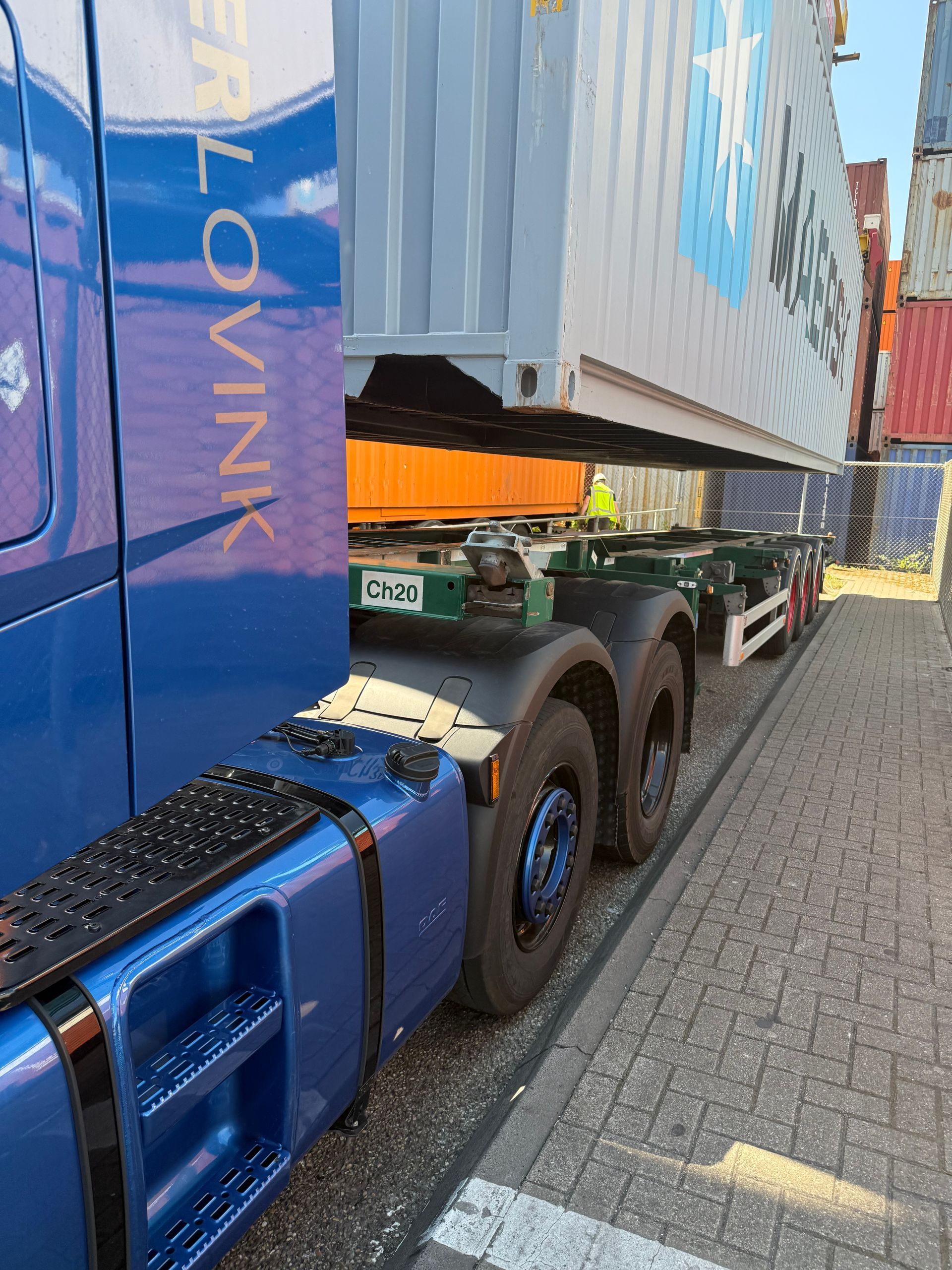 Een blauwe vrachtwagen met oplegger vervoert een witte zeecontainer over een geasfalteerde weg, gezien vanaf de zijkant.