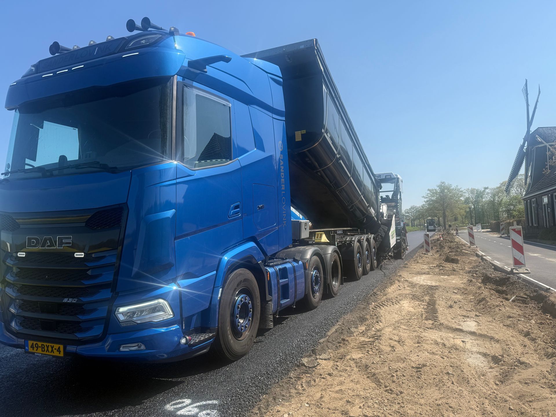 Blauwe kiepwagen op een grindweg naast een bouwterrein, met een verhoogde zwarte aanhanger.