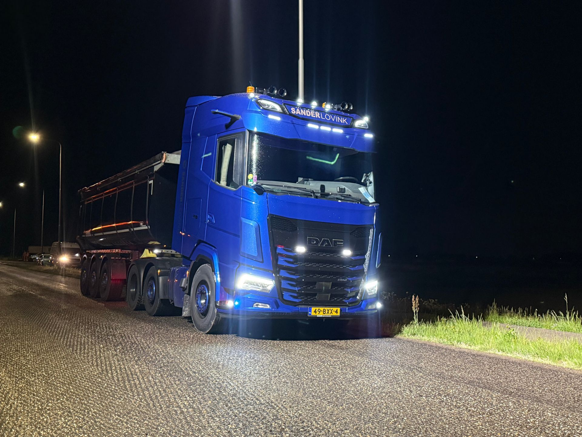 Blauwe vrachtwagen geparkeerd op een onverharde weg 's nachts met koplampen aan.