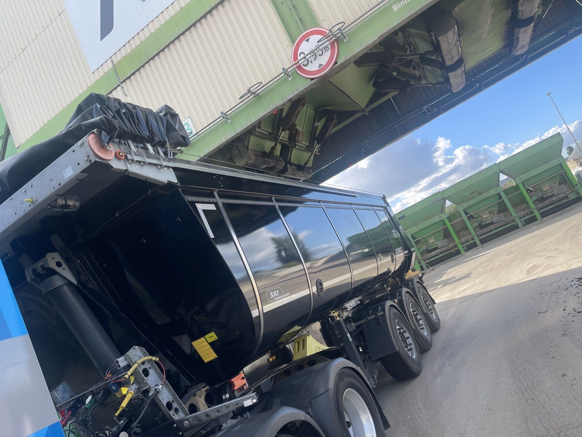 Een zwarte kiepwagen lost lading onder een groene transportband op een industrieterrein onder een blauwe hemel.