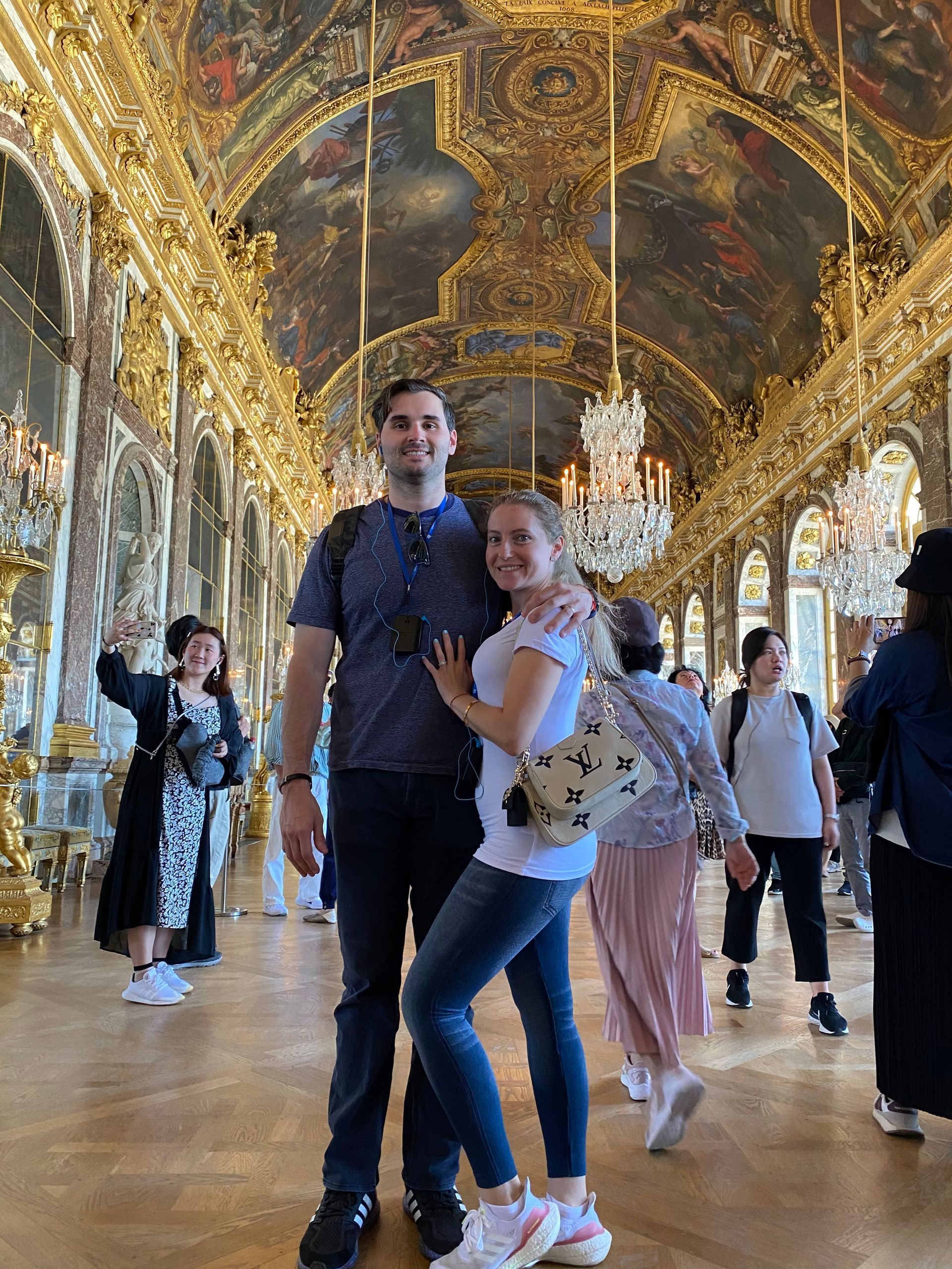 Couple posing in a gilded hall with ornate ceiling, mirrors, and chandeliers; tourists walk in the background
