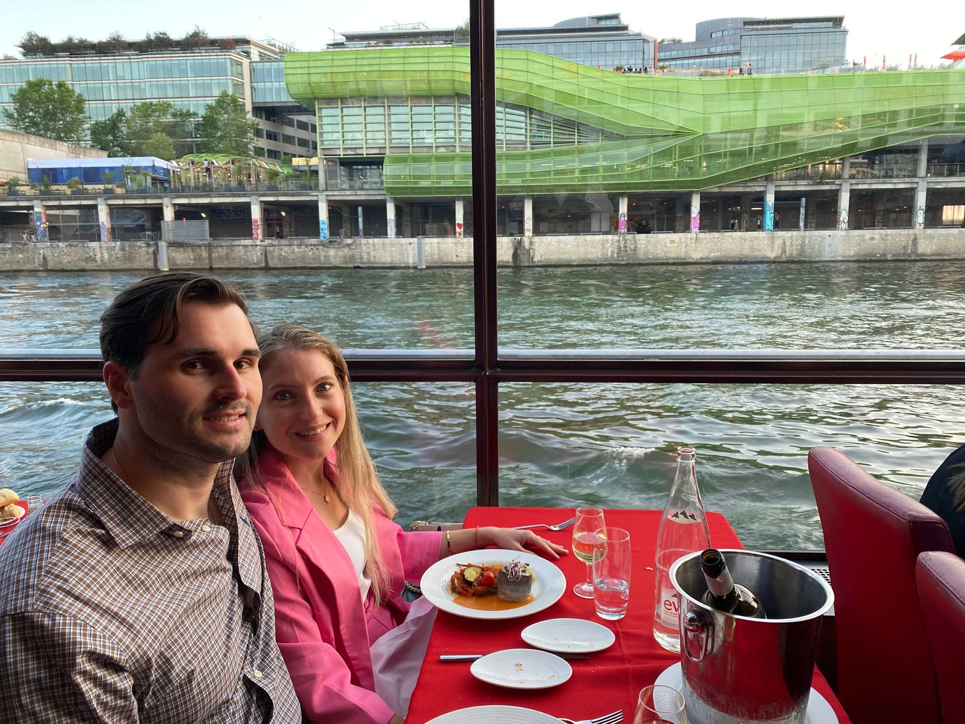 Couple dining by a river, seated at a red table with drinks and plates, city buildings in the background